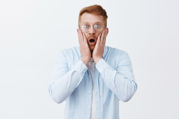 Free photo man being shocked witnessing terrible scene. portrait of stunned terrified redhead male in glasses and shirts dropping jaw, gasping from amazement, staring worried over gray wall