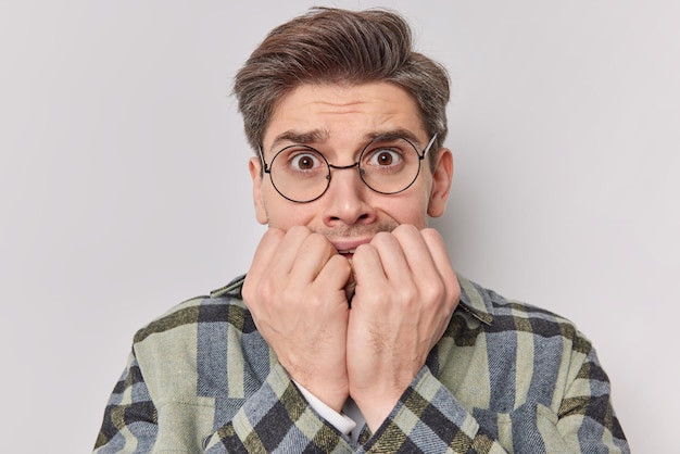 Free photo man bites finger nails looks with nervous wears spectacles and casual clothes poses on white. troubled hipster guy tries to control feelings