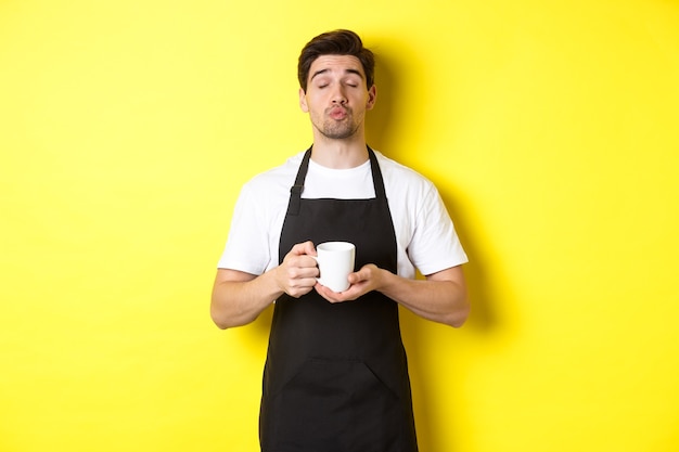 Man in black apron bringing cup of coffee and waiting for kiss, standing over yellow background.