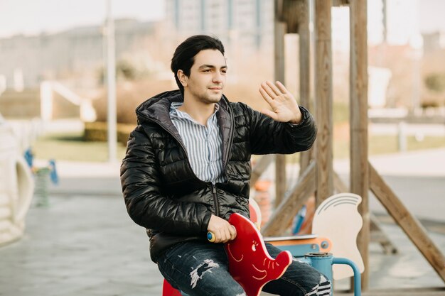 Man in black jacket sitting on the childish carousel in the park and greeting or calling someone with hand gests. High quality photo