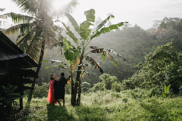 Man in black raincoat touching girlfriend's face on nature. Couple of tourists posing in rain-forest.