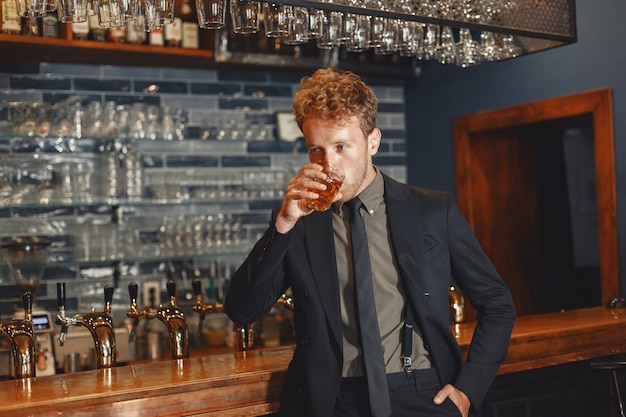 Free photo man in a black suit drinks alcohol. attractive guy drinks whiskey from a glass..