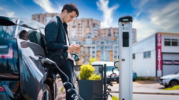 Free photo man charging his electric car at charge station and using smartphone