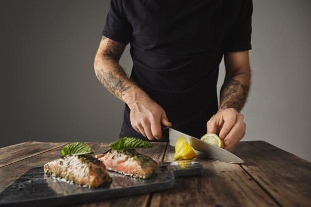 Man cooks healthy meal, cut limon behind decorated with mint leaf two raw pieces of salmon in white wine sause with spices and herbs presented on marble deck prepared for grill