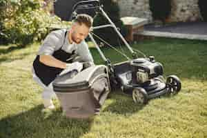 Free photo man cutting grass with lawn mover in the back yard. male in a black apron. guy repairs.