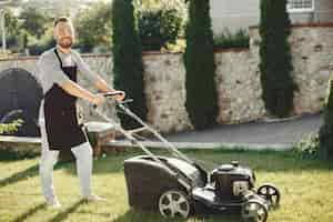 Free photo man cutting grass with lawn mover in the back yard. male in a black apron.