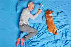 Free photo man dog owner of old age sleeps peacefully together with pet poses in bed wears pajama and socks lying on soft pillow sees sweet dreams. mature bearded man rests at bedroom. people slumber concept