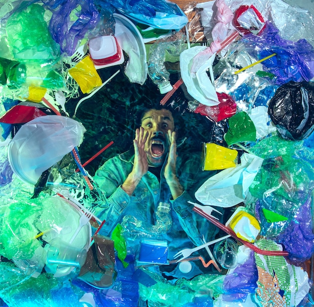 Man drowning in water under plastic recipients pile, garbage. Used bottles and packs filling world ocean killing people.