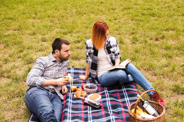 Free photo man eating banana looking at her girlfriend reading book at the picnic