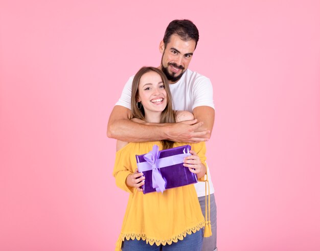 Man embracing her girlfriend holding purple gift box over pink background