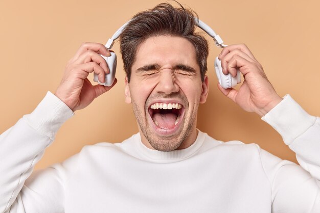 man exclaims loudly keeps mouth wide opened takes off stereo headphones listens music with high volume dressed in casual white jumper isolated on beige.