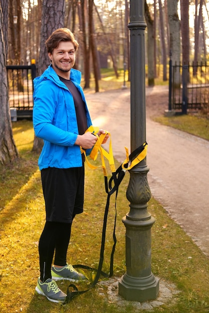 Man fitness model in a blue sport jacket doing workouts with fitness trx strips in a park.