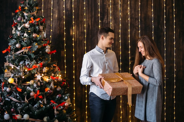 Man gives a present to the woman standing before a Christmas tree