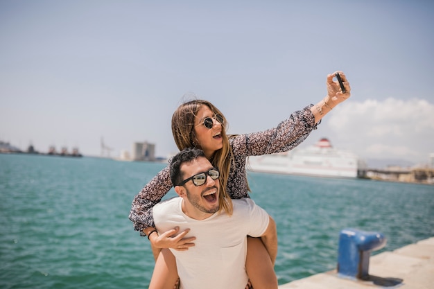 Man giving her girlfriend piggyback ride taking selfie on cell phone near the sea