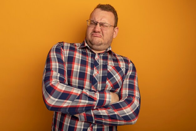 Man in glasses and checked shirt  dissatisfied with frowning face with crossed arms standing over orange wall