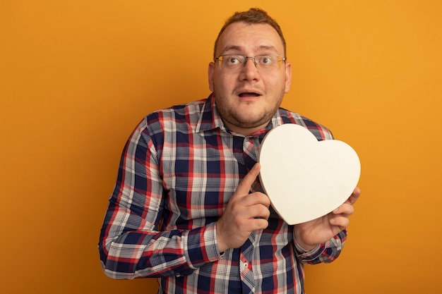 Free photo man in glasses and checked shirt holding cardborad heart looking aside confused standing over orange wall