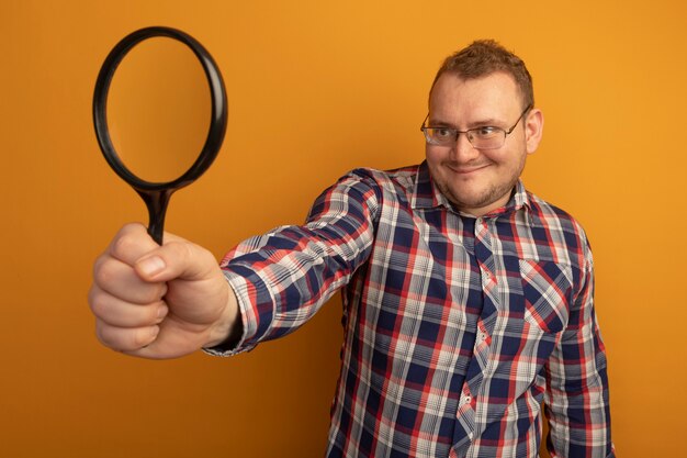 Man in glasses and checked shirt holding magnifying glass looking at it with smile on face standing over orange wall