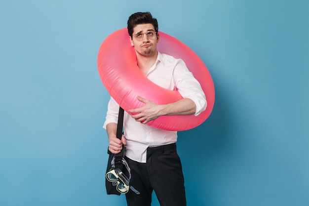 Free photo man in glasses holds inflatable circle, diving mask and bag for documents. guy in office clothes posing on blue space.