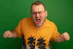 Free photo man in glasses and orange shirt shouting with clenches fists angry and frustrated standing over green wall