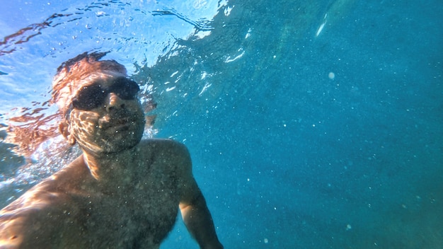 Man in goggles swimming under the blue and transparent water of the Mediterranean sea. Holding the camera