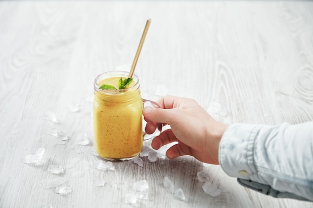 Free photo man hand takes transparent rustic jar with tasty healthly freshly made yellow smoothie from mango