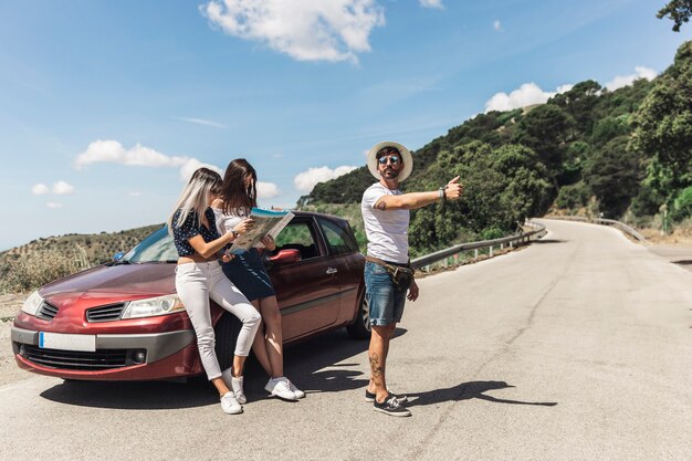 Man hitchhiking on road standing in front of female friends looking at map