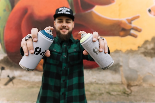 Free photo man holding aerosol cans with hands standing against graffiti wall