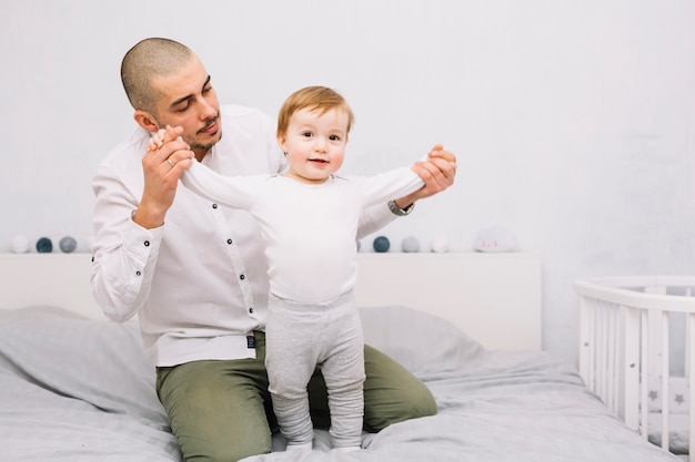 Free Photo man holding hands of funny little baby standing on bed