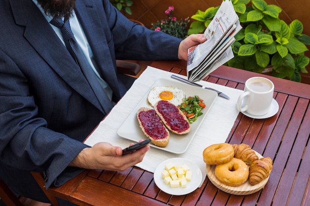 Free photo man holding smartphone and newspaper during breakfast