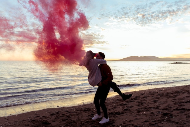 Free photo man holding woman in arms with pink smoke bomb on sea shore