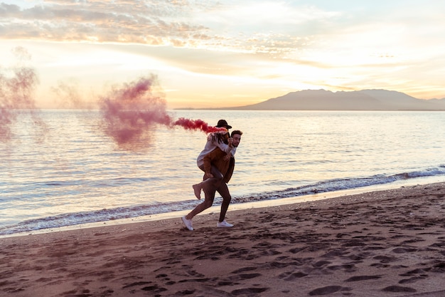 Man holding woman with smoke bomb on back on sea shore 