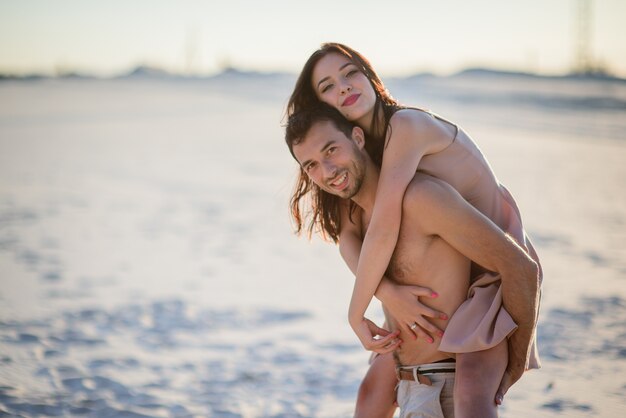 Man holds pretty woman on his back walking on the beach