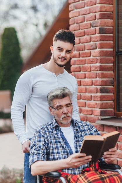 A man is carrying his father near nursing home, they are having fun and they are laughing while reading a book