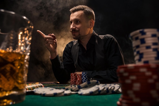 Free photo man is playing poker with a cigar and a whiskey. a man winning all the chips on the table with thick cigarette smoke. the concept of victory. glass with whiskey and stack of chips in the foreground