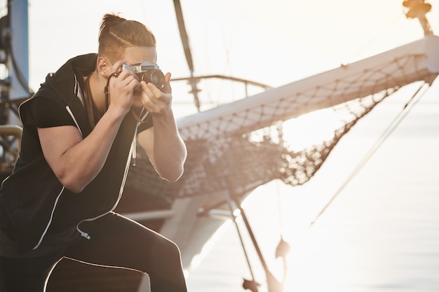 Free photo man is searching for best shots. focused photographer during work standing near yacht bending while looking through camera, taking pictures of sea or harbour, making shots with lifestyle concept