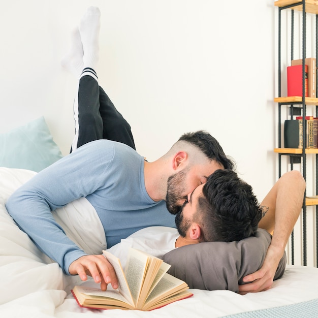 Man kissing his boyfriend lying on bed with le's leaning on wall