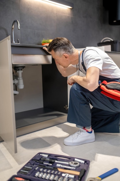 Free photo man kneeling down to inspect pipes under sink