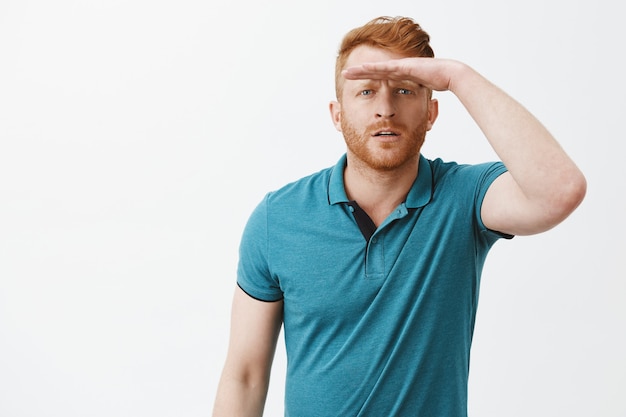 Free photo man looking far away, squinting and holding palm on forehead to cover eyes from sunlight and see clearly, standing focused and interested in greet polo shirt over grey wall