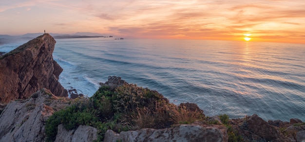 A man looks out to sea from a cliff at sunset.