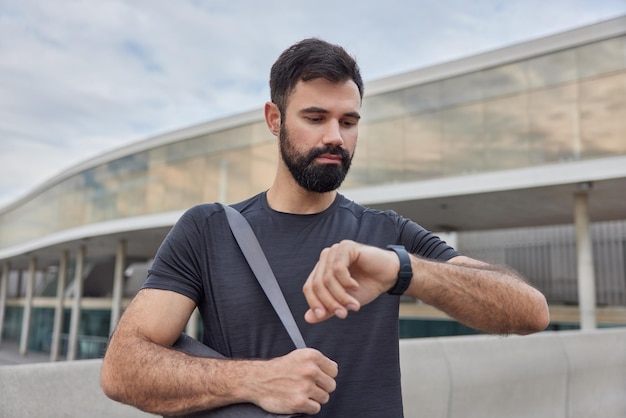 man looks at smartwatch checks time or burned calories after workout carries rolled karemat practices yoga outside wears casual black t shirt poses outdoors. Sport in city