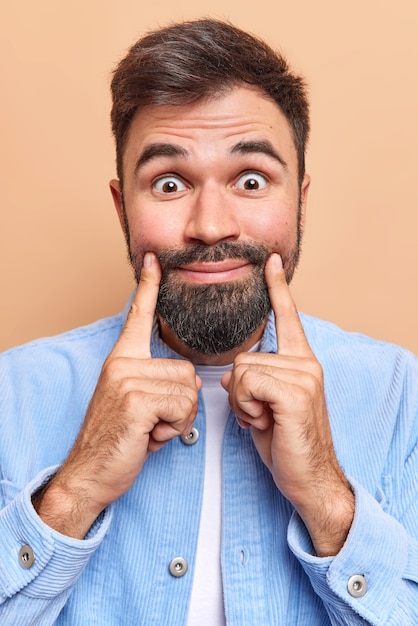 Free photo man looks surprisingly at camera keeps index fingers near lip corners forces smile dressed in blue velvet shirt isolated on brown