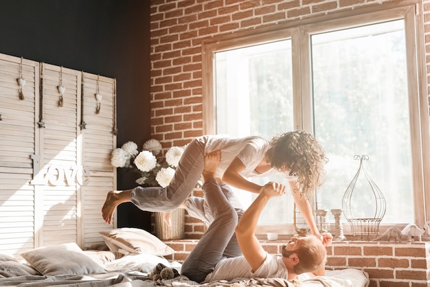 Free photo man lying on bed carrying her wife on his feet near the window