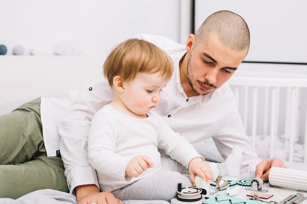 Free photo man lying near little baby with toy sitting on bed near crib