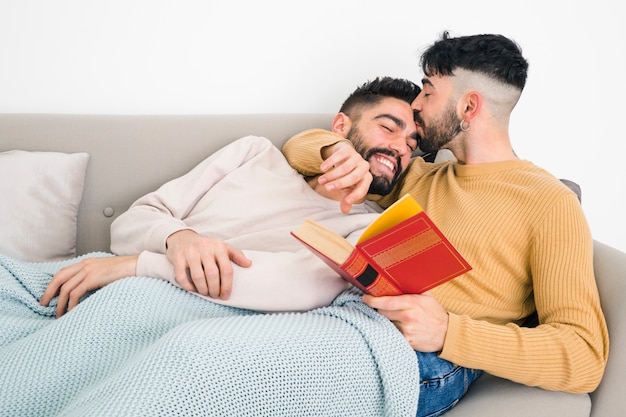 Free photo man lying on sofa kissing his boyfriend's forehead holding book in hand