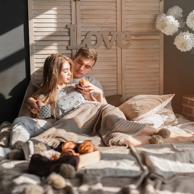 Man lying with her girlfriend feeding croissant to her on bed