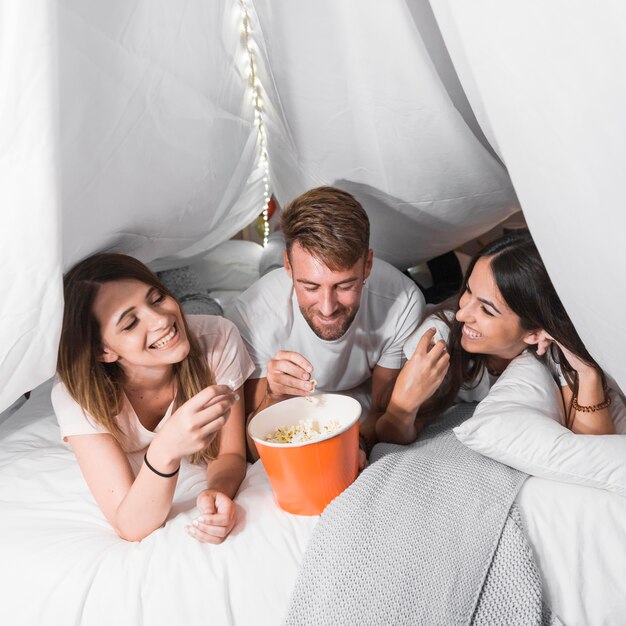 Man lying with two female friends on bed eating popcorns