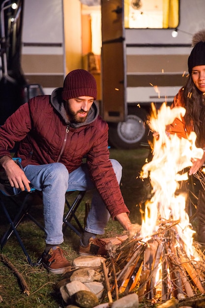 Free photo man making the camp fire stronger in a cold night of autumn in the mountains. tourists with retro camper van.