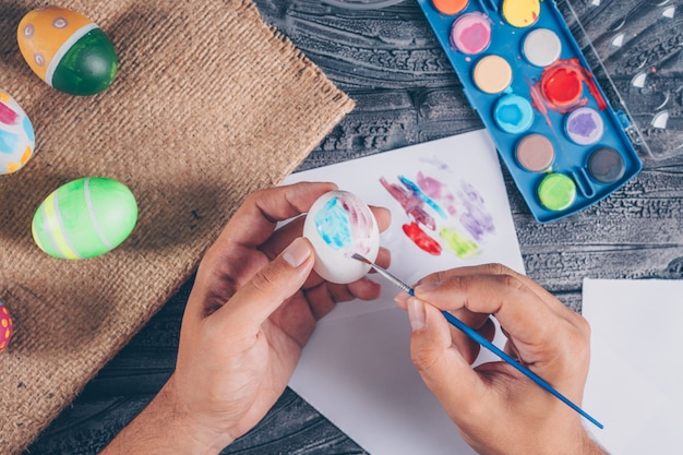 man making an easter egg with paint on dark wooden background top view