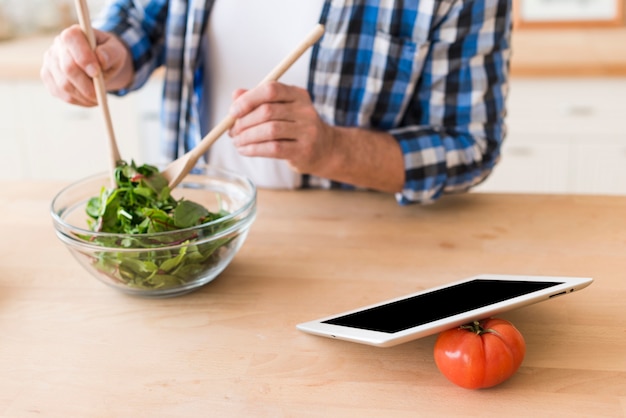 Free Photo man mixing the leafy vegetable with wooden tablespoon in the glass bowl with digital tablet and tomato on wooden desk