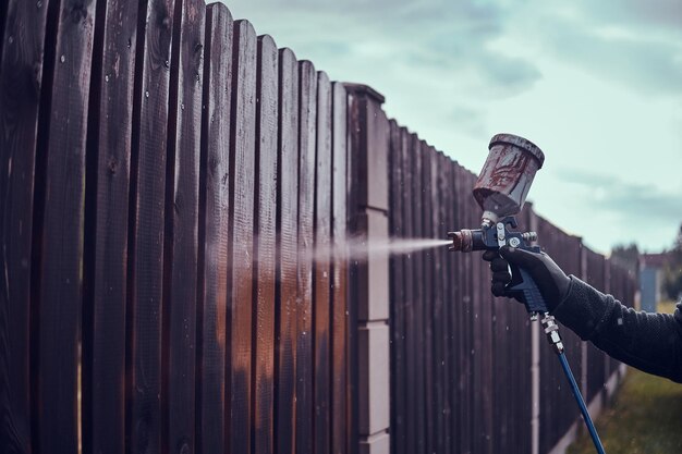 Man n protective gloves is applying paint to the old fence using airbrush at his garden.